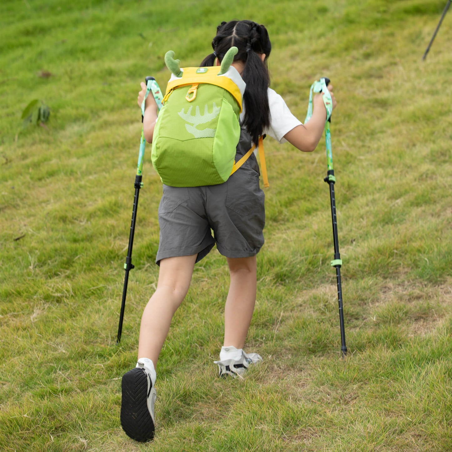 mädchen mit grünem kinderrucksack wandert mit trekkingstöcken auf wiese
